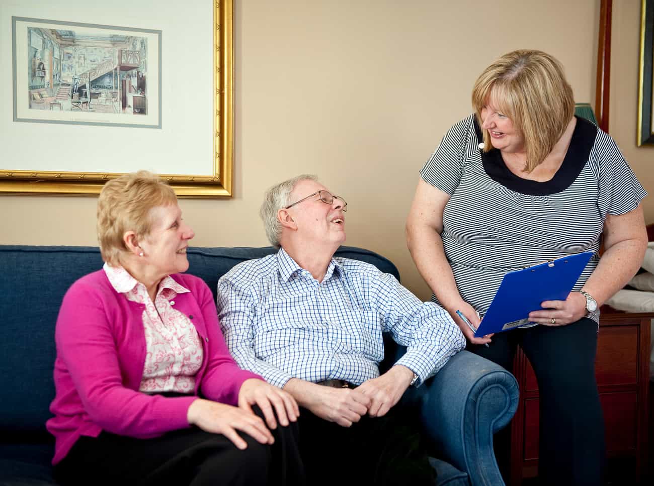 An older woman and man sitting on a sofa with a carer standing beside them and talking with them.