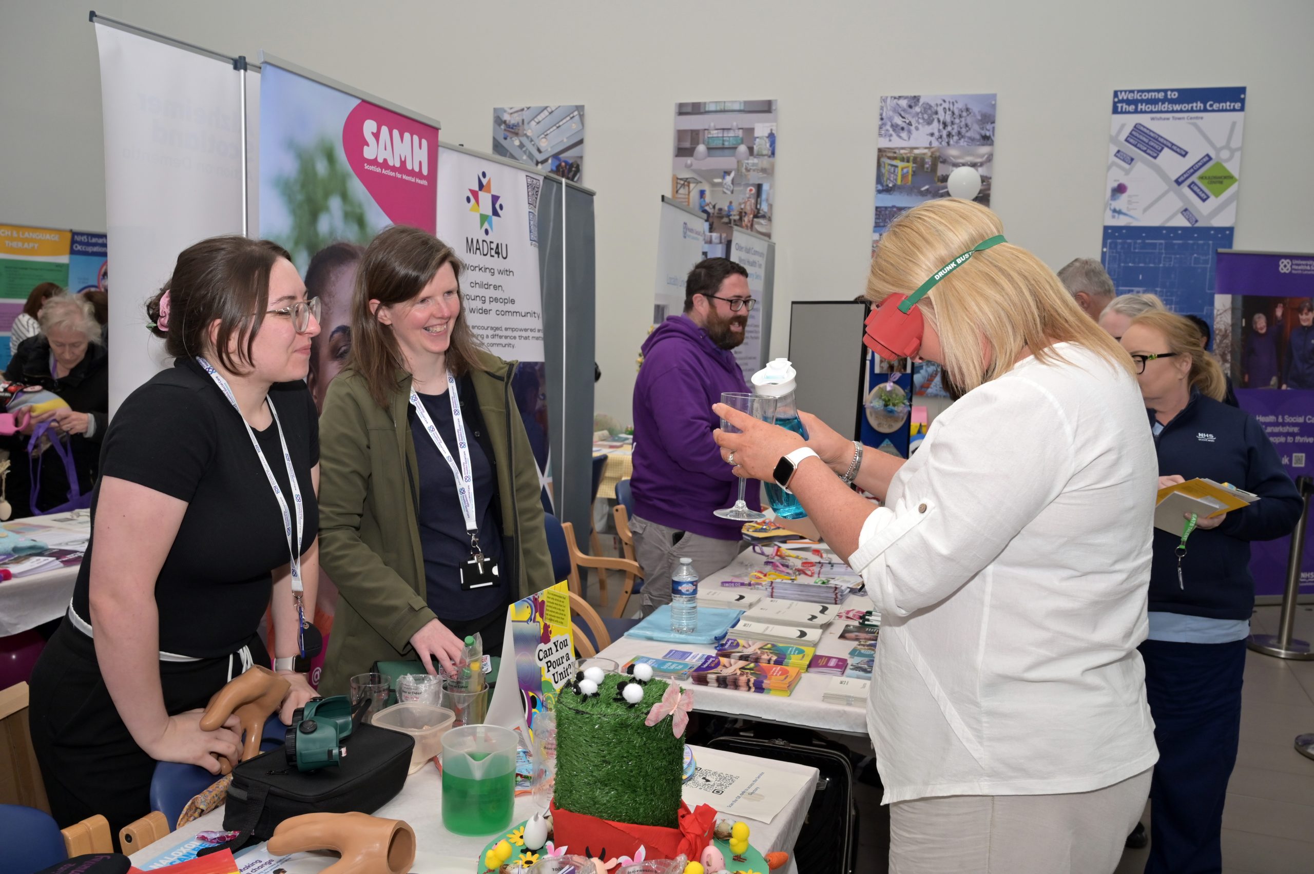 Community members engaging with staff and volunteers at an indoor health improvement event, with tables displaying wellbeing resources and information as part of a local initiative to empower residents in Motherwell.
