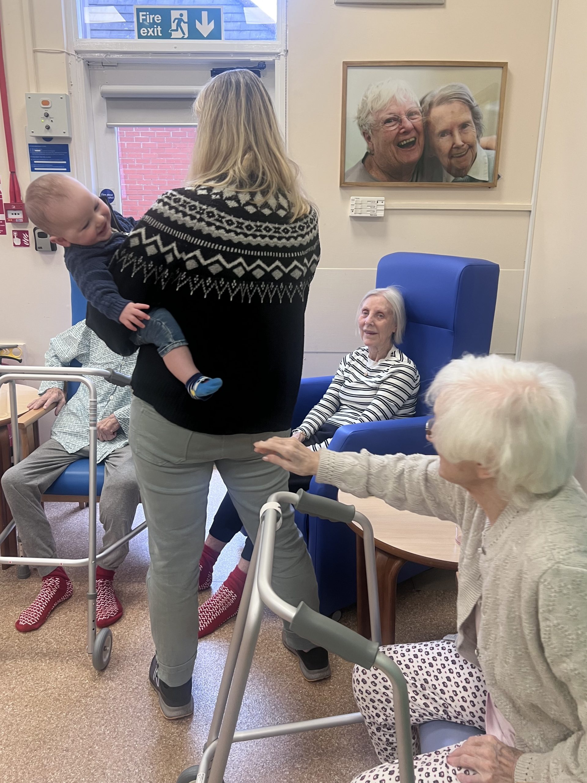 Mother holding her baby and showing the child to three elderly people seated nearby, sharing a warm moment as part of an intergenerational breastfeeding initiative in a community setting.